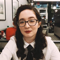 Emi, a light-skinned person with dark curled hair in a 1940s style, smiles at the camera like they have a secret. They're wearing dark tortoiseshell cat-eye glasses, dark red lipstick, and a white collared shirt with dark tie.